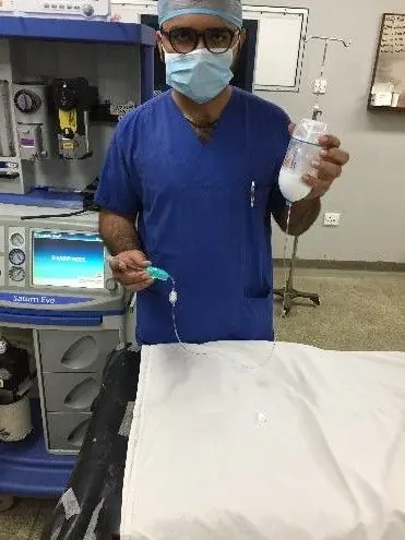 A clinician in theatre scrubs demonstrating an infusion product beside an anaesthesia workstation